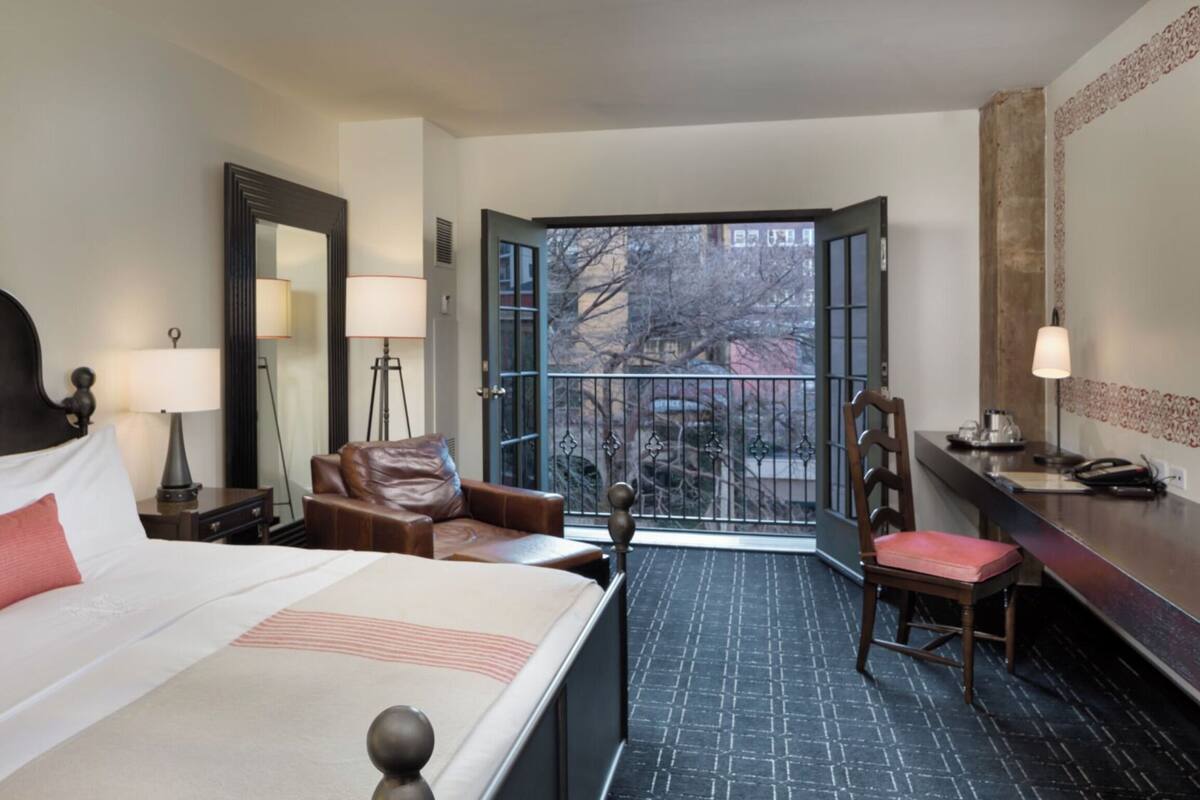 Een hotelkamer met een bed, bureau, stoel en een balkon met uitzicht op bomen en gebouwen.