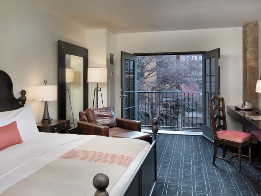 A hotel room with a bed, a desk, a chair, and a balcony with a view of trees and buildings.