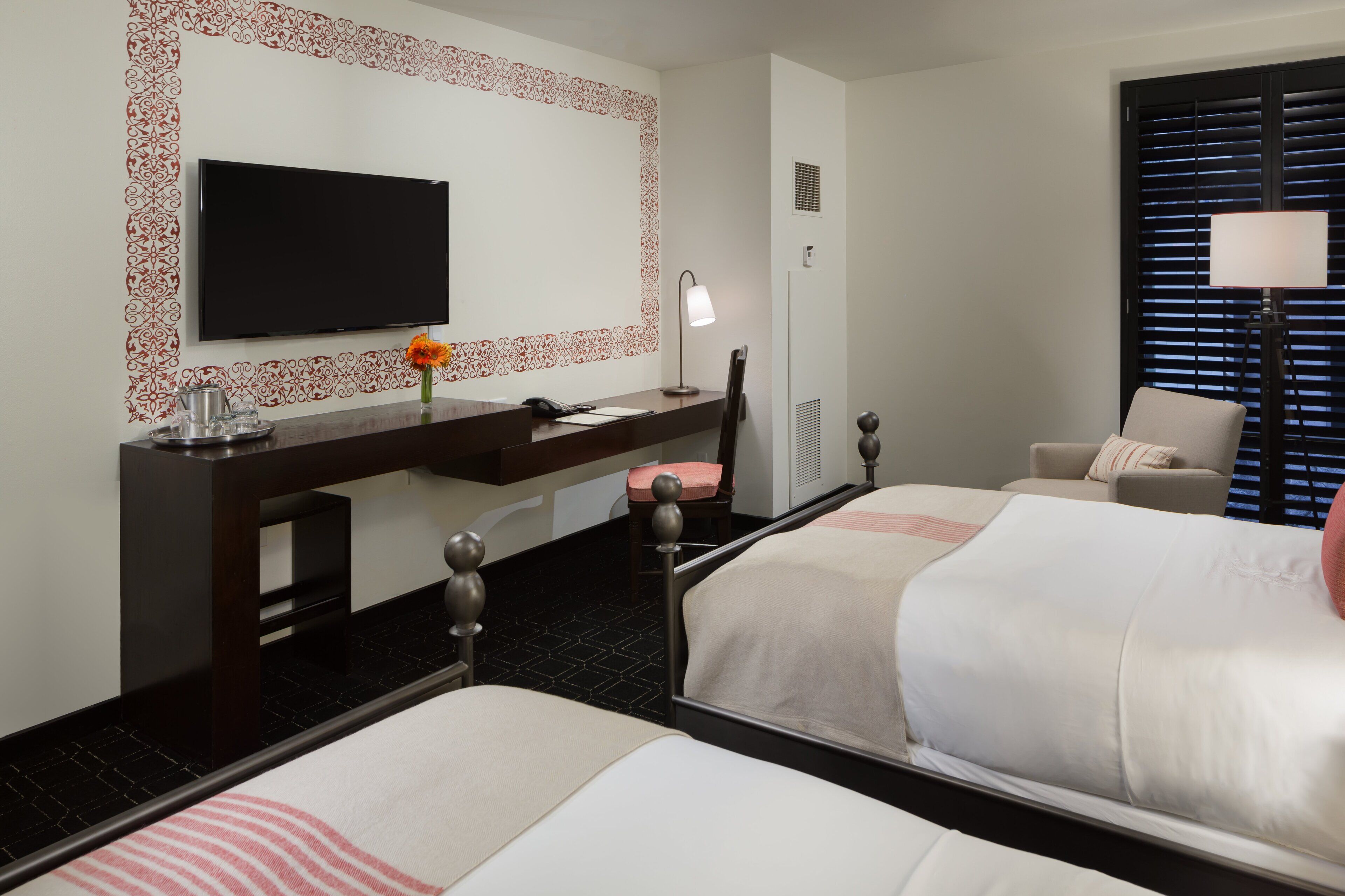 Une chambre d’hôtel avec deux lits, une télévision, un bureau avec une lampe, une chaise et une fenêtre avec des stores.