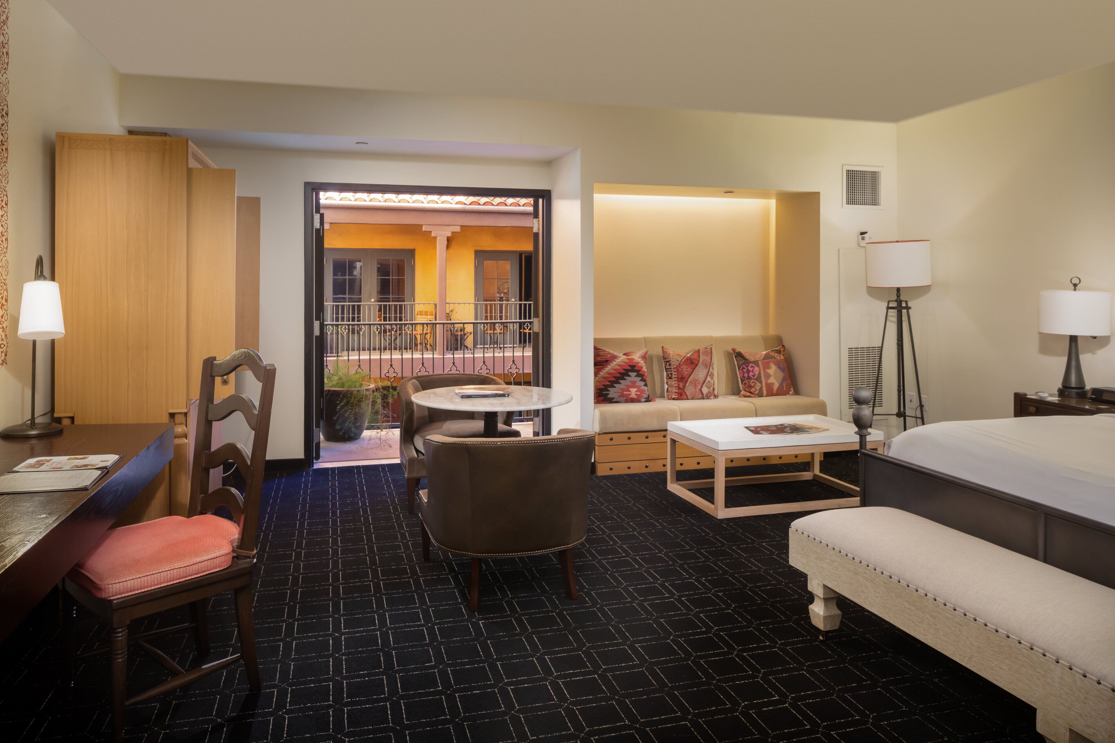 A hotel room with a bed, a sofa, a dining table with chairs, a small round table, and a lamp.