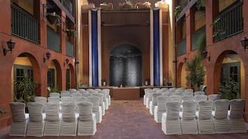 Un espacio interior con filas de sillas blancas dispuestas para un evento, una fuente decorativa en el centro y suelo de ladrillo.