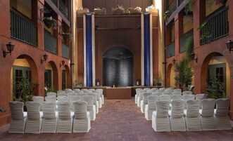 Un espacio interior con filas de sillas blancas dispuestas para un evento, una fuente decorativa en el centro y suelo de ladrillo.