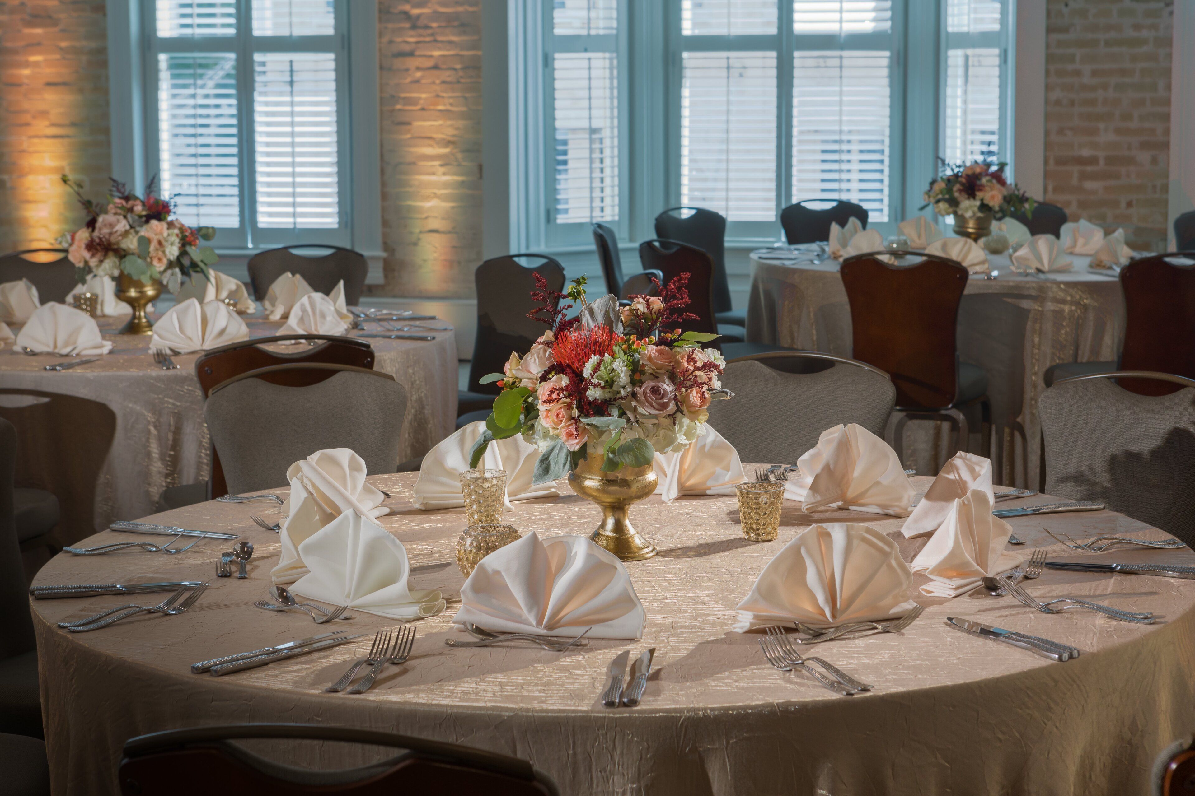 A dining area with round tables set for a formal event, featuring white tablecloths, folded napkins, and floral centerpieces.