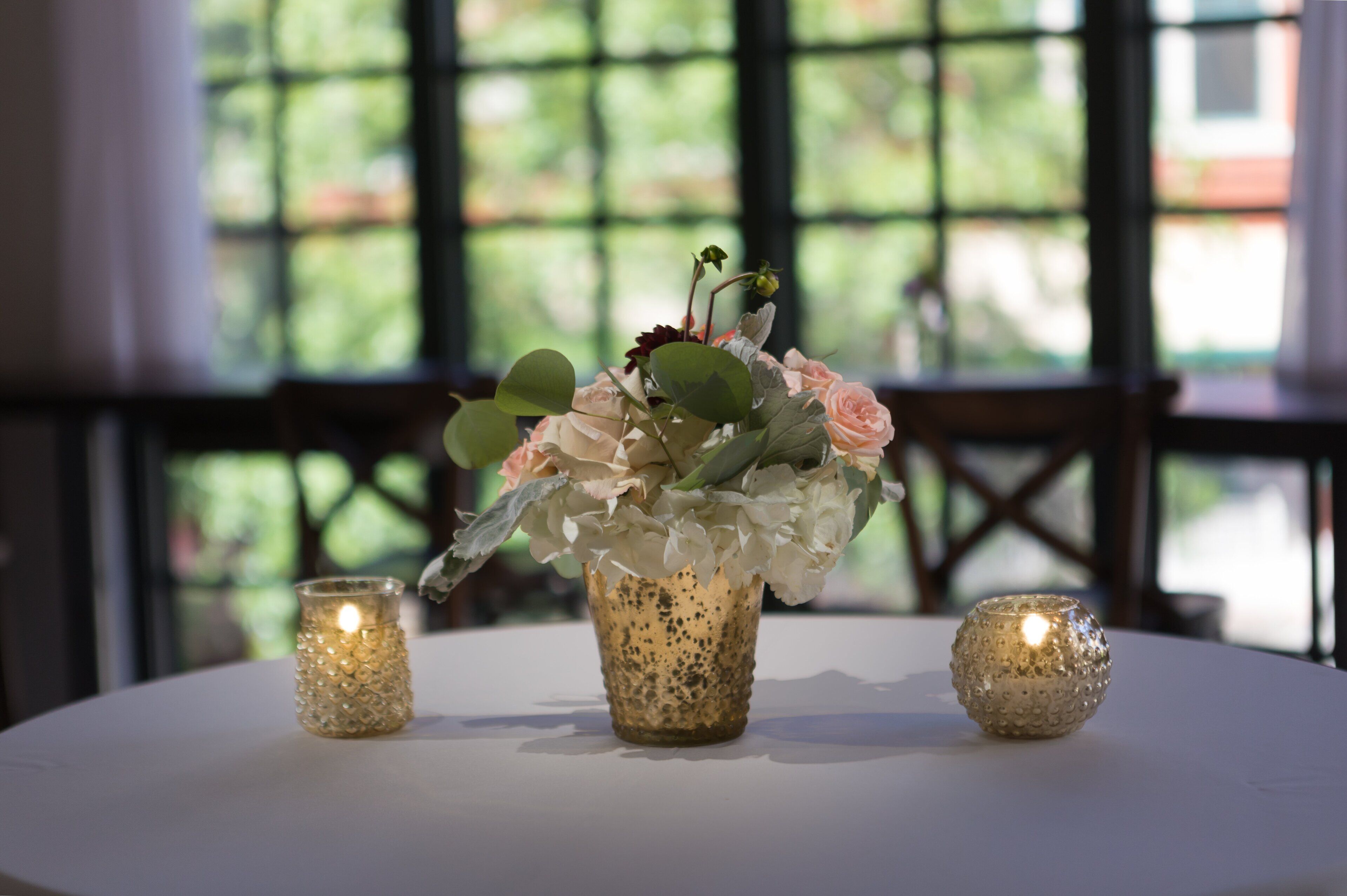 A table with a floral arrangement in a textured vase, two lit candles, and a view of greenery through a window.