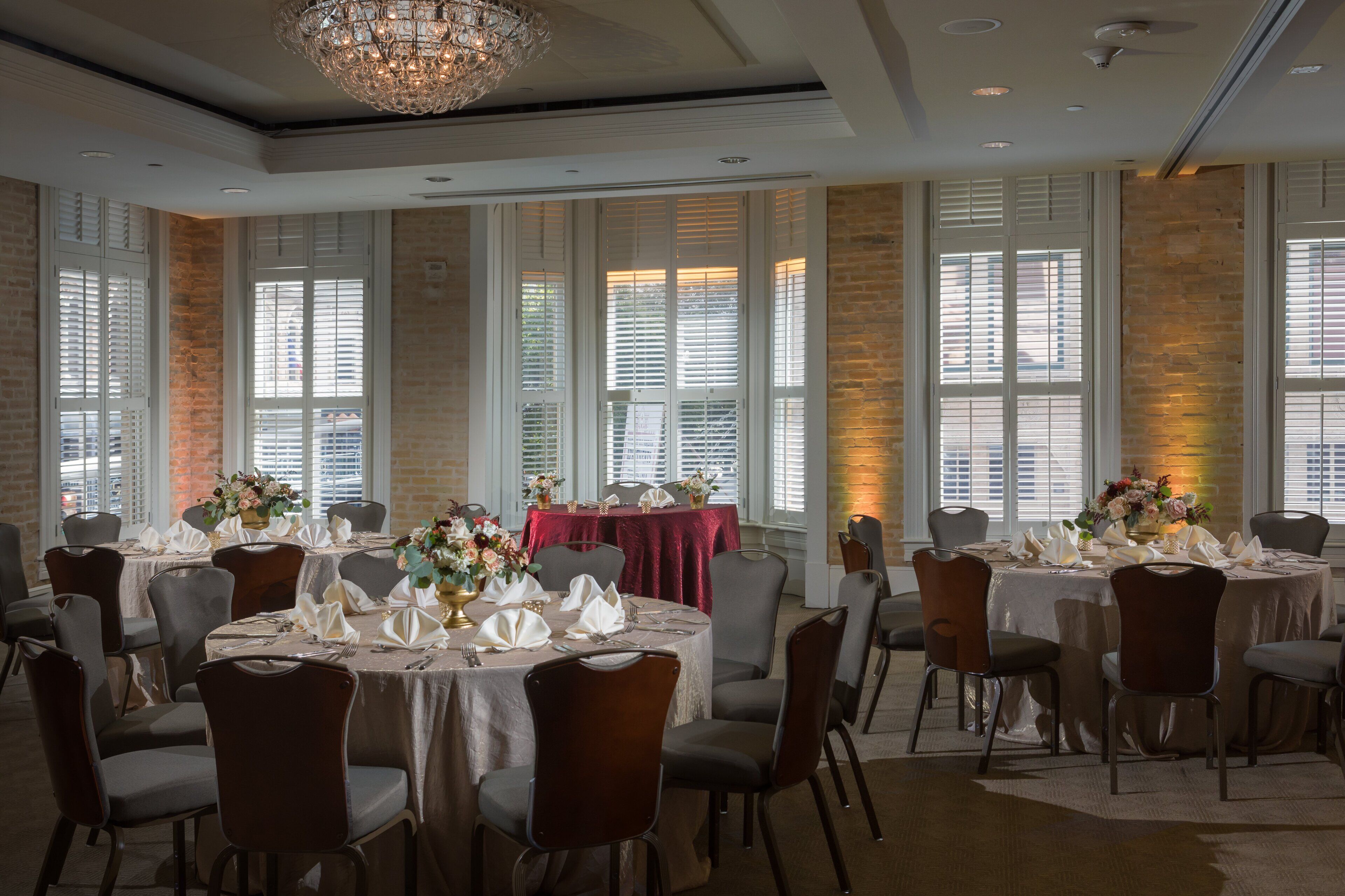 A banquet hall with round tables set for dining, large windows with blinds, and a chandelier.