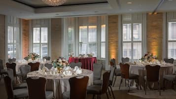 A banquet hall with round tables set for dining, large windows with blinds, and a chandelier.