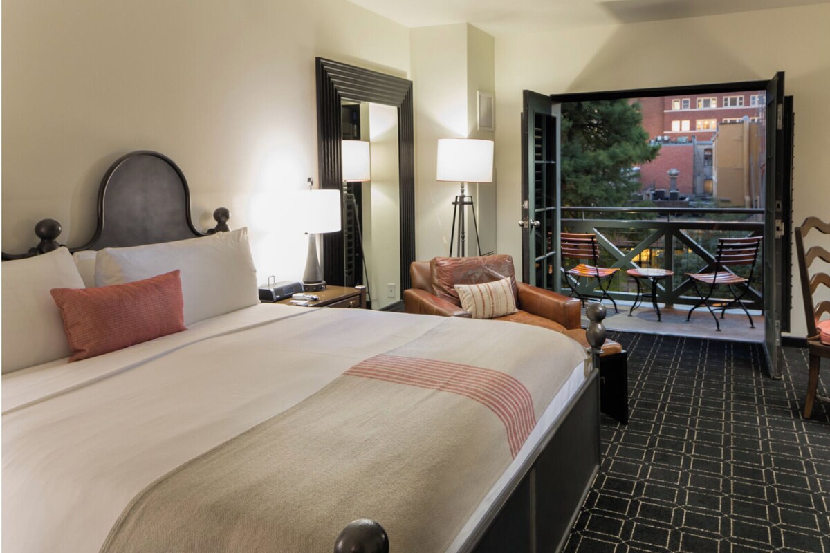 Habitación de hotel con una cama grande, un escritorio con silla, un balcón con sillas y vistas a edificios.