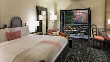 A hotel room with a large bed, a desk with a chair, a balcony with chairs, and a view of buildings outside.