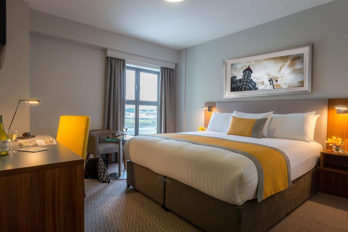 A hotel room with a bed, desk, chair, and a painting on the wall.