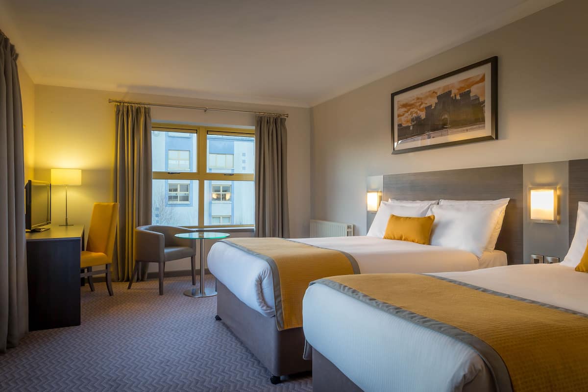 A hotel room with two beds, a desk, a chair, a window with curtains, and a painting on the wall.