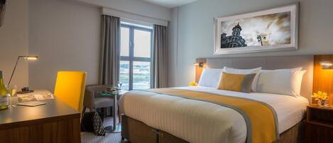 A hotel room with a large bed, a desk with a lamp, a chair, a small table with a drink, and a painting on the wall.