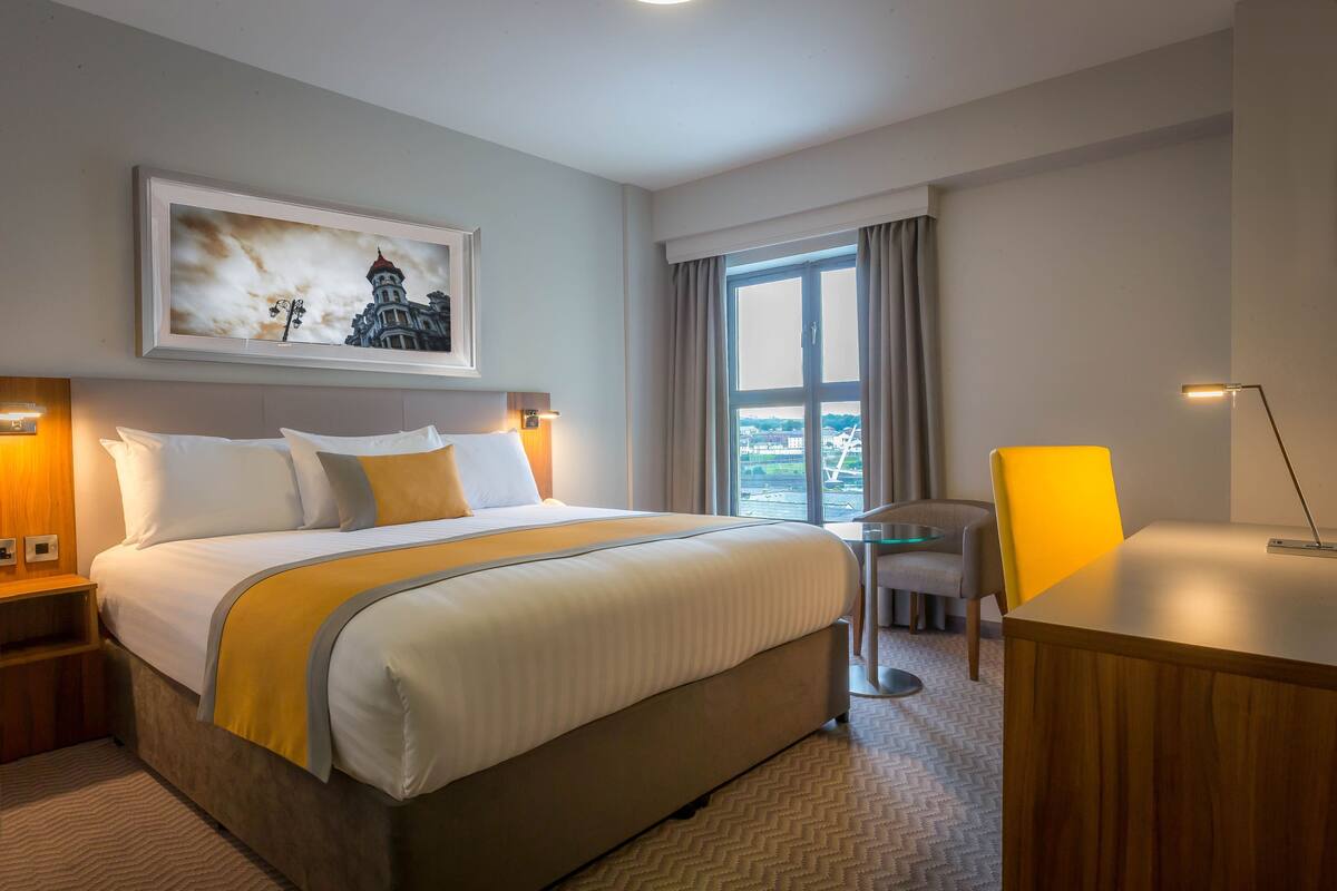 A hotel room with a large bed, a desk, a chair, a window with curtains, and a framed picture on the wall.