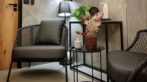 Een moderne kamer met een houten deur, twee grijs gestoffeerde fauteuils, een klein rond tafeltje met een bloevas, een lamp en een wandlamp.