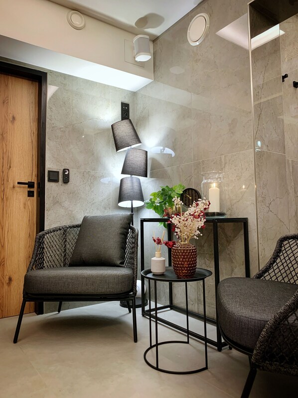 Une pièce moderne avec une porte en bois, deux fauteuils gris à revêtement en tissu, une petite table ronde ornée d’un vase de fleurs, une lampe et un luminaire fixé au mur.