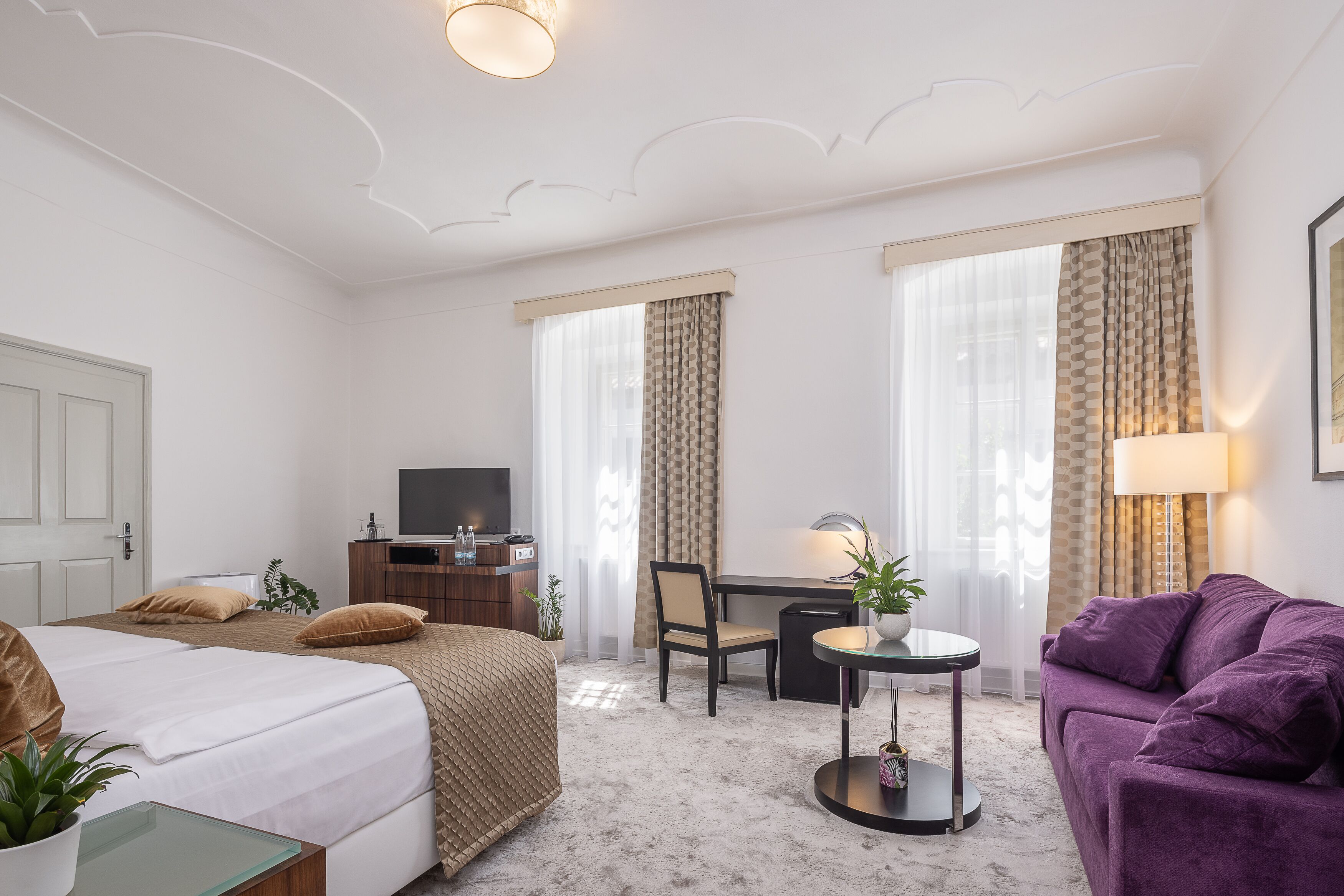 Ein Hotelzimmer mit einem Bett, einem Schreibtisch, einem Sessel und einem violetten Sofa, sowie einem kleinen Tisch mit einer Lampe.