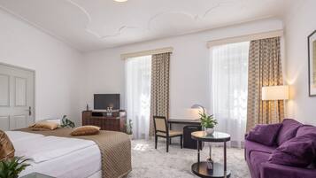 Ein Hotelzimmer mit einem Bett, einem Schreibtisch, einem Sessel und einem violetten Sofa, sowie einem kleinen Tisch mit einer Lampe.