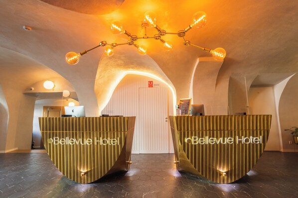 Un área de recepción con dos mostradores identificados como "Bellevue Hotel", una lámpara de araña moderna y un suelo con patrón hexagonal.