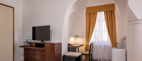 Habitación de hotel compacta con tocador de madera, televisor, pequeña mesa con lámpara, silla y electrodoméstico blanco.