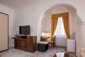Habitación de hotel compacta con tocador de madera, televisor, pequeña mesa con lámpara, silla y electrodoméstico blanco.