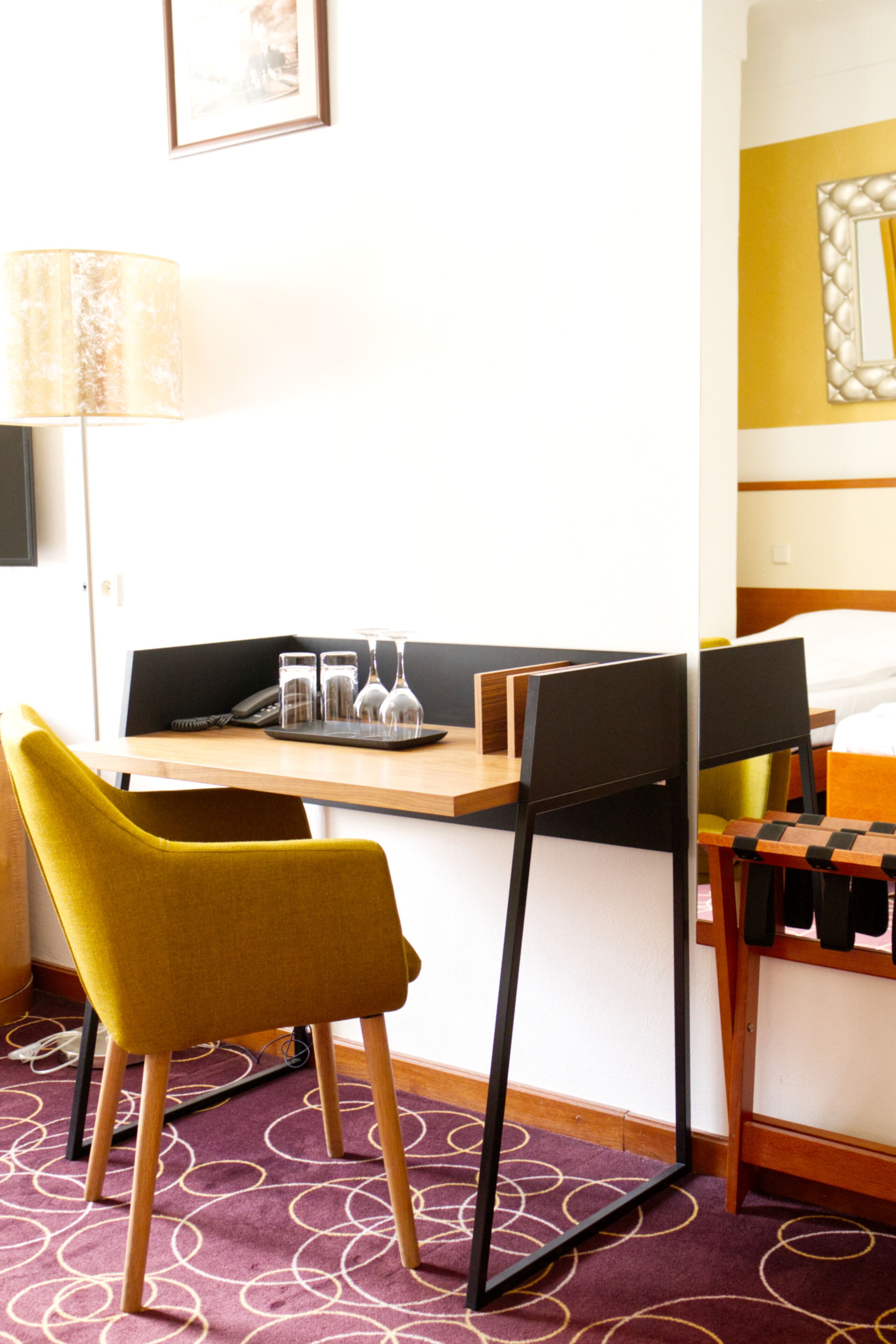 A hotel room with a desk, two chairs, a bed, a lamp, and a small table with glasses and a phone.
