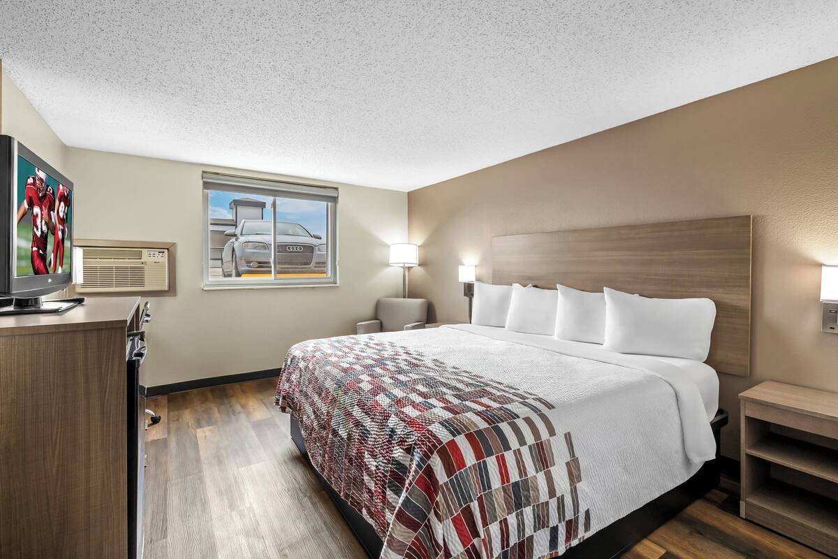 A hotel room with a large bed, a TV on a wooden dresser, a window with a view of a car, and a wall-mounted heater.