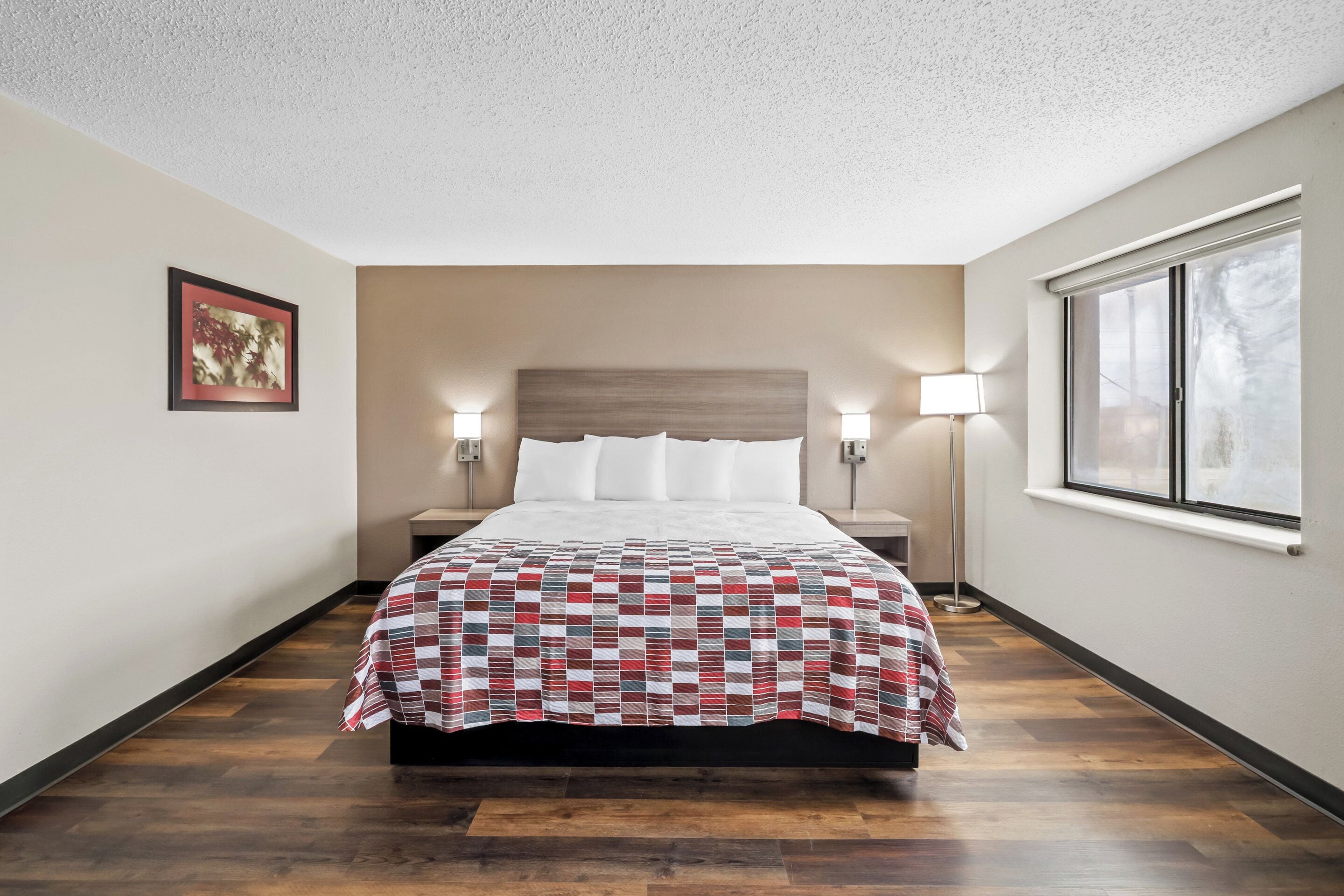 A hotel room with a wooden floor, a bed with a checkered bedspread, two bedside lamps, a nightstand, a framed picture on the wall, and a window with a view.