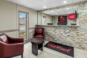A Red Roof hotel reception area with a stone wall, a Red Roof sign, a digital display for free Wi-Fi, a reception desk, a chair, and a small table.