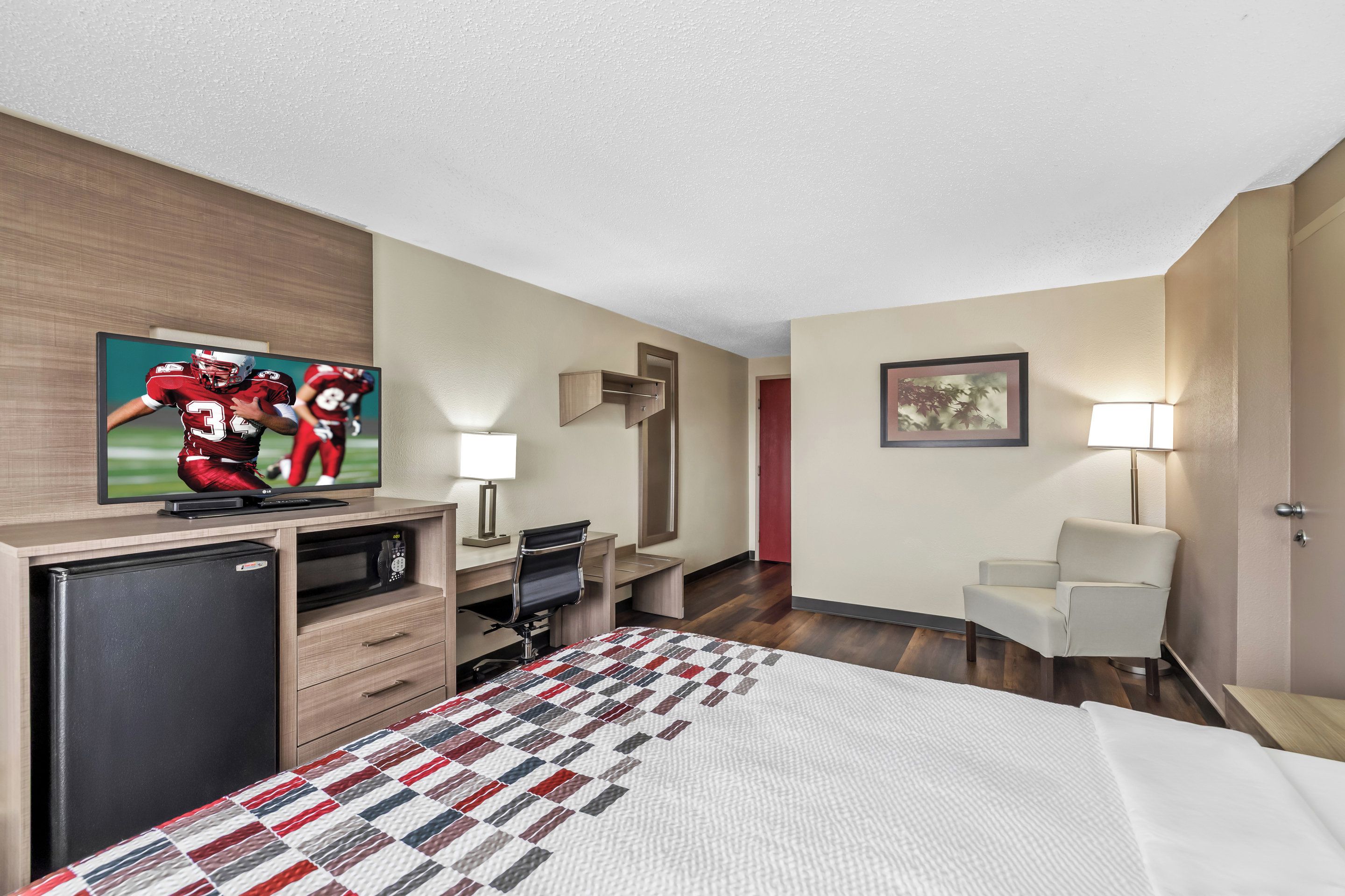 A hotel room with a bed, a desk with a computer, a television, a refrigerator, a chair, and a painting on the wall.