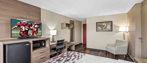 A hotel room with a bed, a desk with a computer, a television, a refrigerator, a chair, and a painting on the wall.
