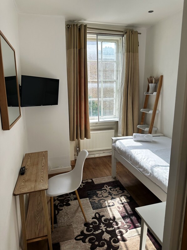 A small, well-lit room with a bed, a desk, a chair, a TV, and a window with curtains. - Somerset Hotel Baker Street London (London)
