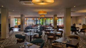 A spacious restaurant with high ceilings, large windows, and a patterned carpet. Tables are neatly arranged with chairs, and there are hanging lamps providing warm lighting. - Cedar Court Wakefield (Wakefield)