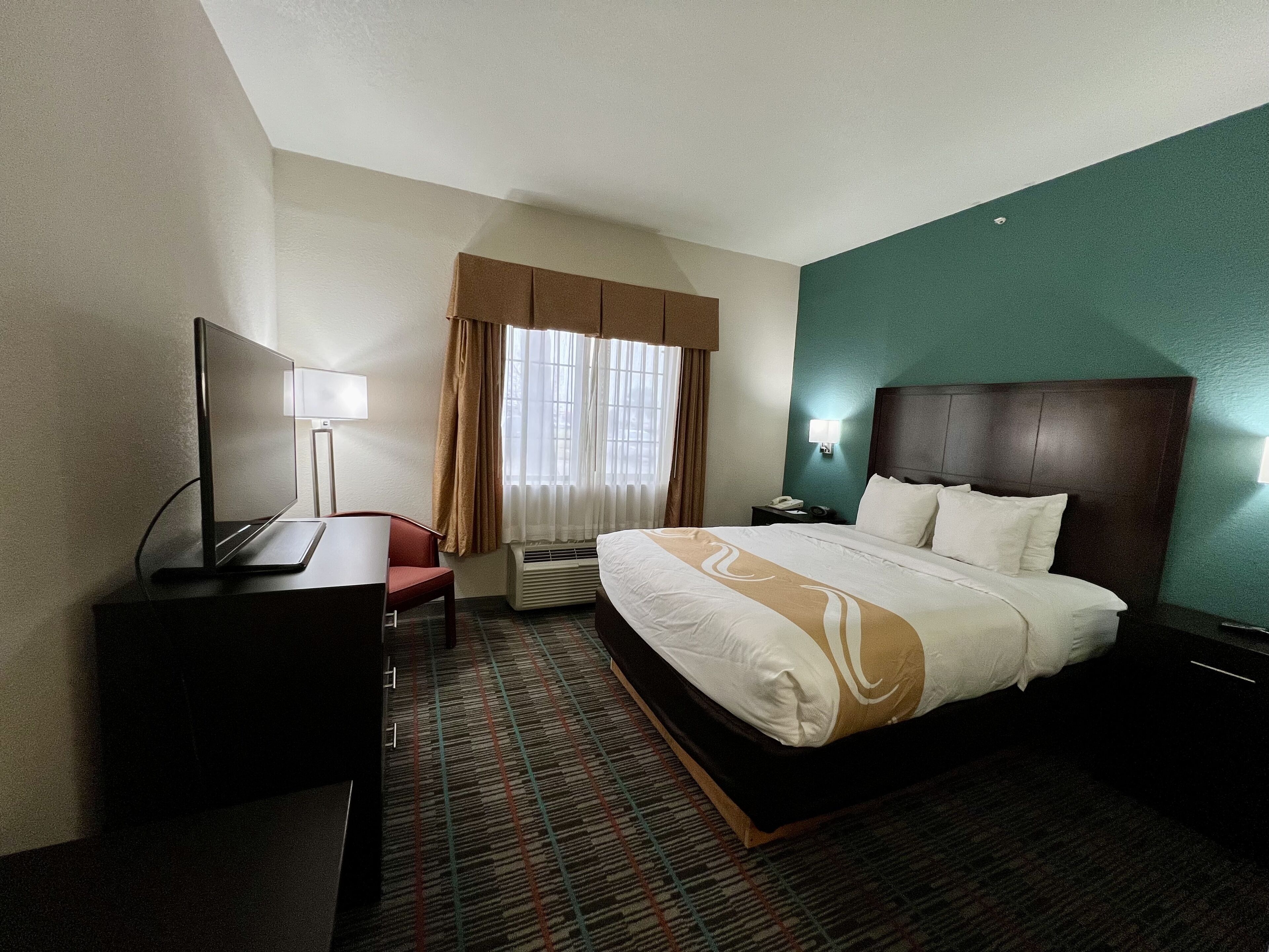 A hotel room with a bed, a desk, a chair, a TV, a lamp, and a window with curtains.