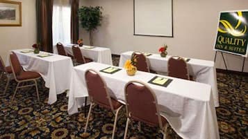 Un salón de conferencias con mesas dispuestas para reuniones, una pantalla de proyector y un cartel de Quality Inn & Suites.