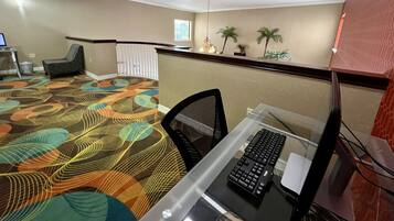Habitación de hotel con escritorio, silla y una alfombra colorida. Se ve una ventana con vistas a palmeras y una planta colgante.