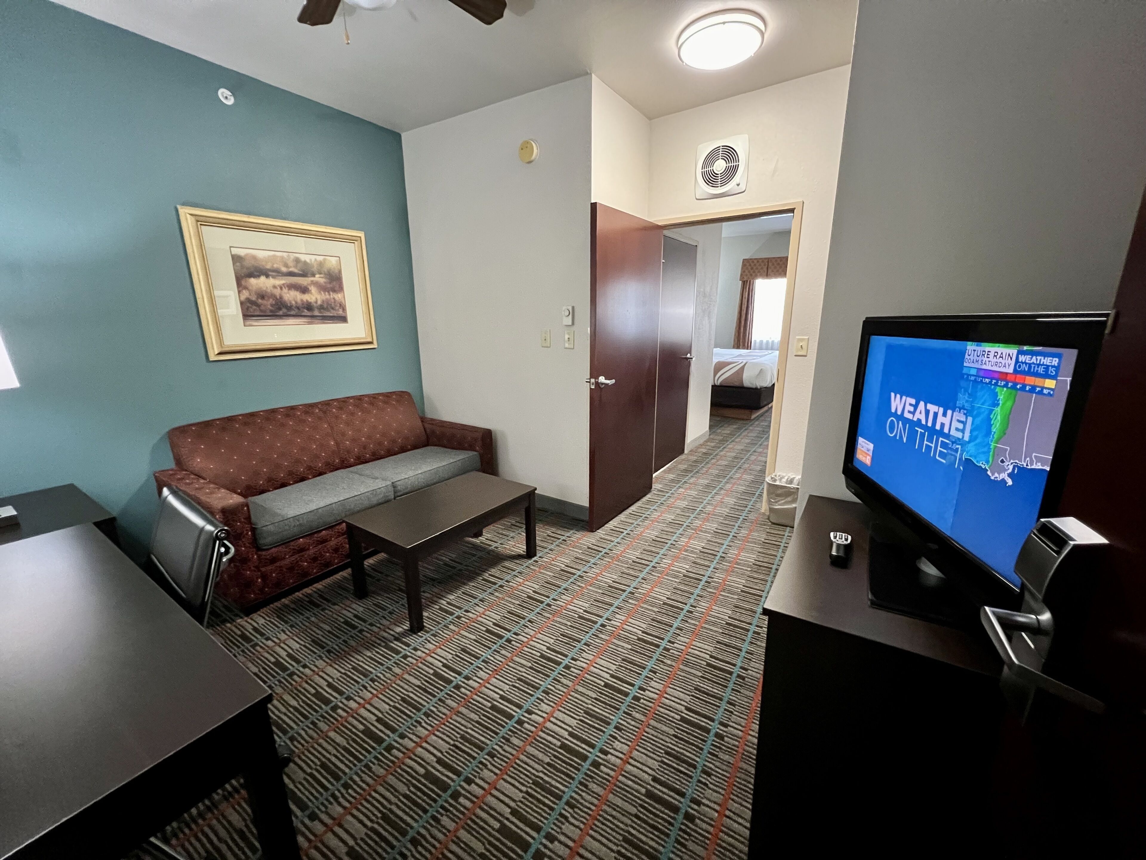 A hotel room with a sofa, a small table, a television displaying weather information, and a patterned carpet.