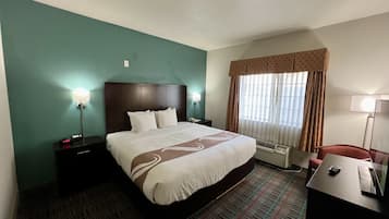 Quarto de hotel com uma cama grande, uma escrivaninha, uma cadeira, uma mesa de cabeceira, um abajur e uma janela com cortinas.