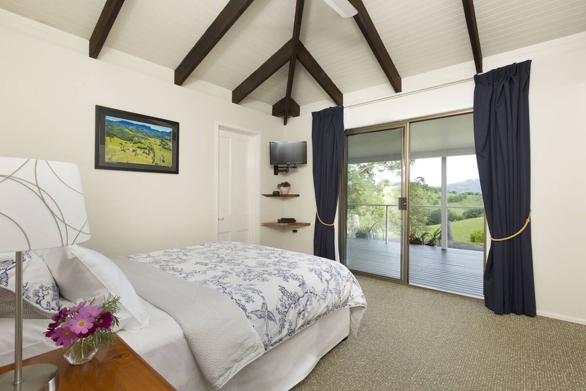 A bedroom with a bed, a TV, a window with curtains, a painting, and a view of greenery.