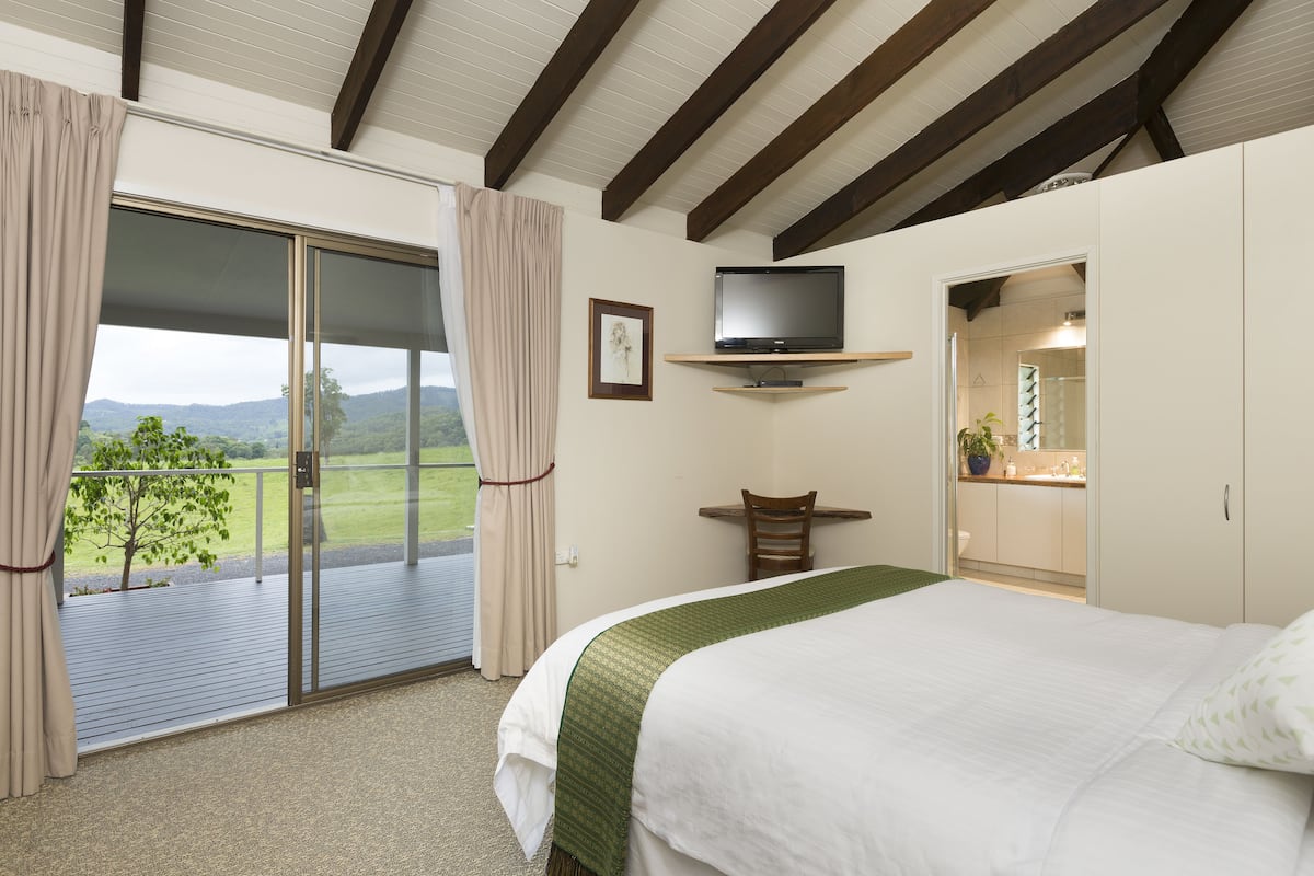 A bedroom with a large bed, a television mounted on a shelf, a desk, and a sliding glass door leading to a balcony with a view of the countryside.
