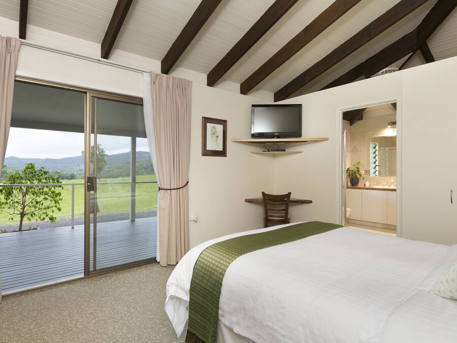 Un dormitorio con una cama grande, un televisor montado en una estantería, un escritorio y un ventanal corredera que da a un balcón con vistas al campo.