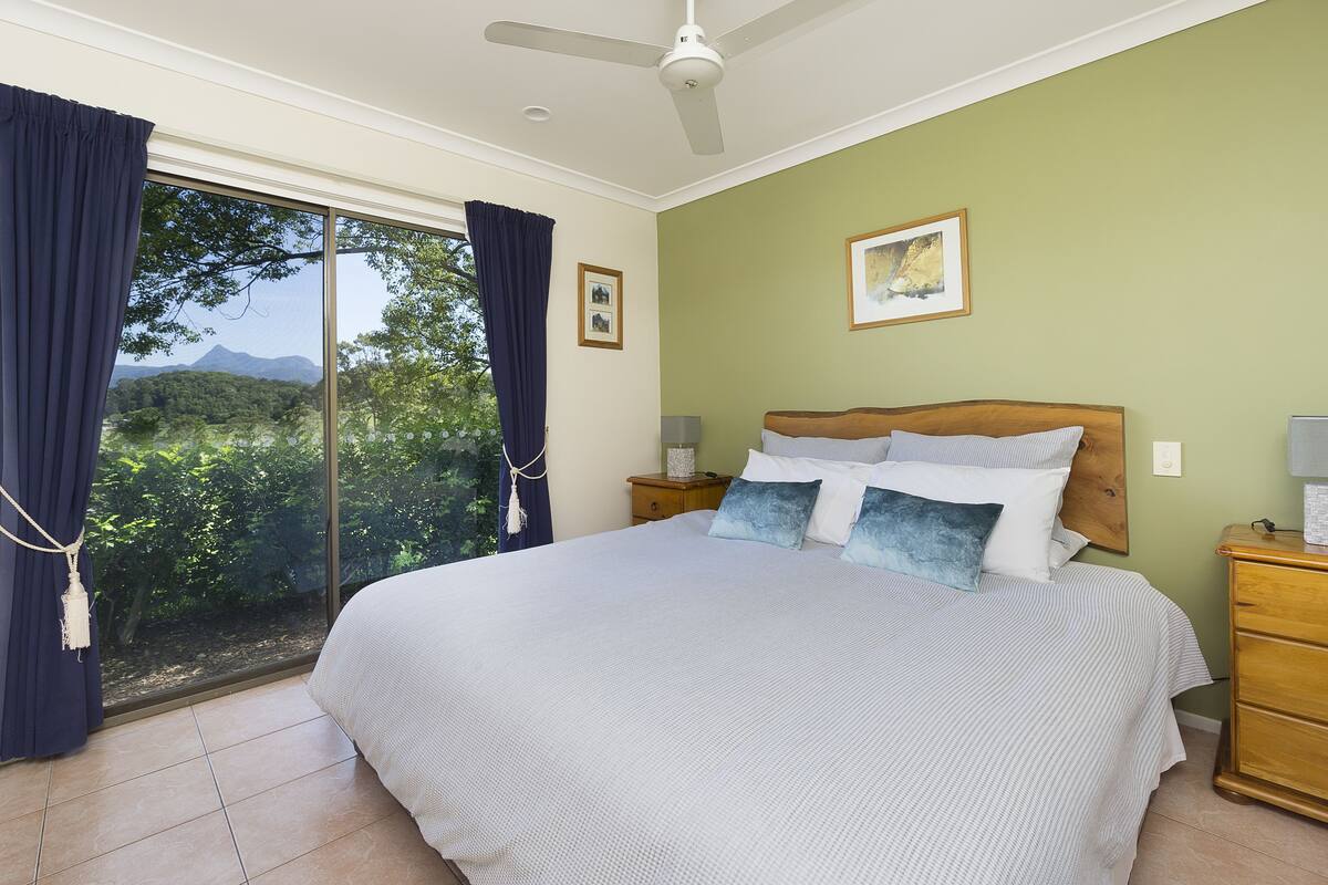 A bedroom with a large bed, a nightstand, a wooden dresser, a ceiling fan, and a window with curtains.