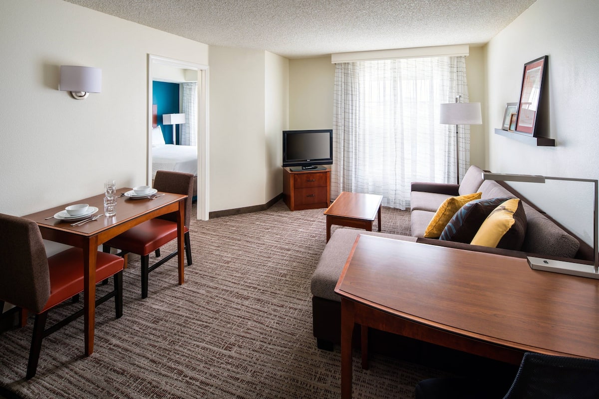 A hotel room with a dining table, chairs, a sofa, a TV, and a window with curtains.