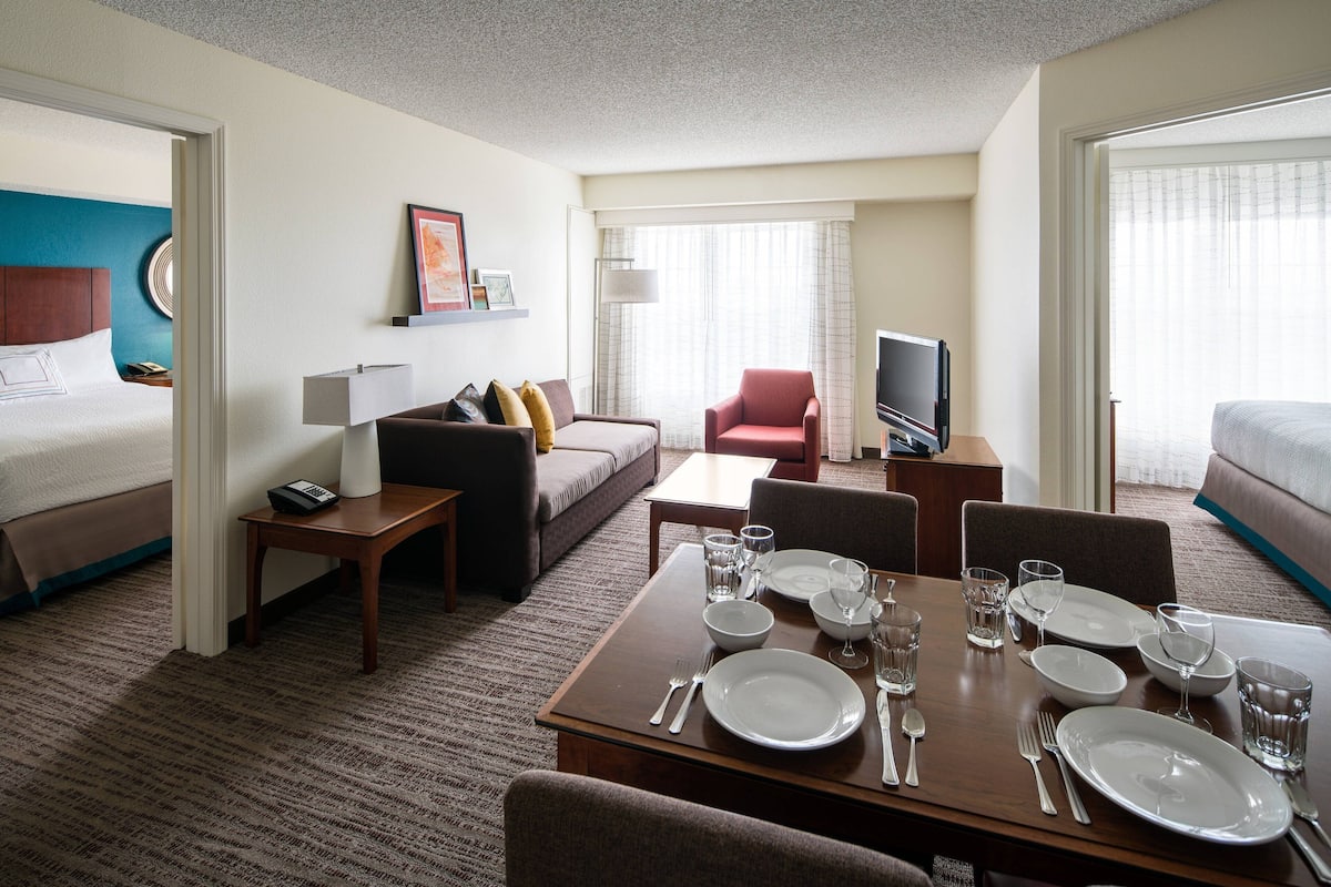 A hotel room with a bed, a sofa, a dining table set with plates and glasses, a TV, and a window with curtains.