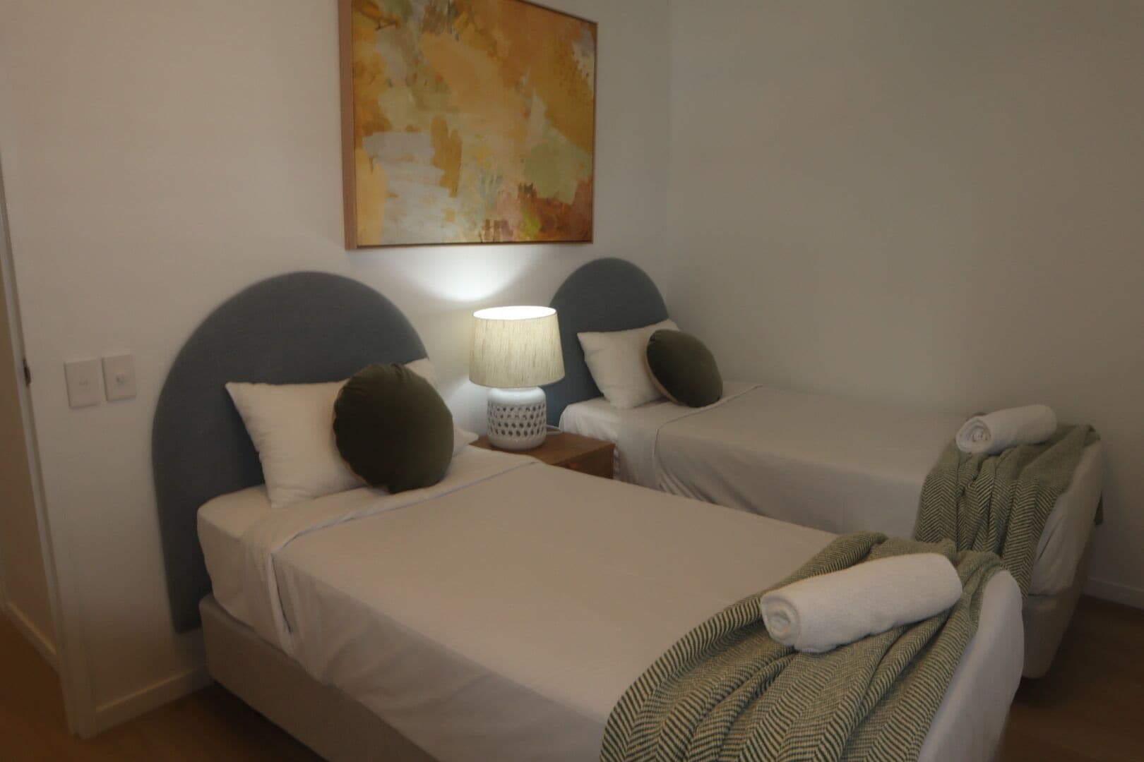 A hotel room with two single beds, each with a headboard, a bedside table with a lamp, and a blanket on one bed.