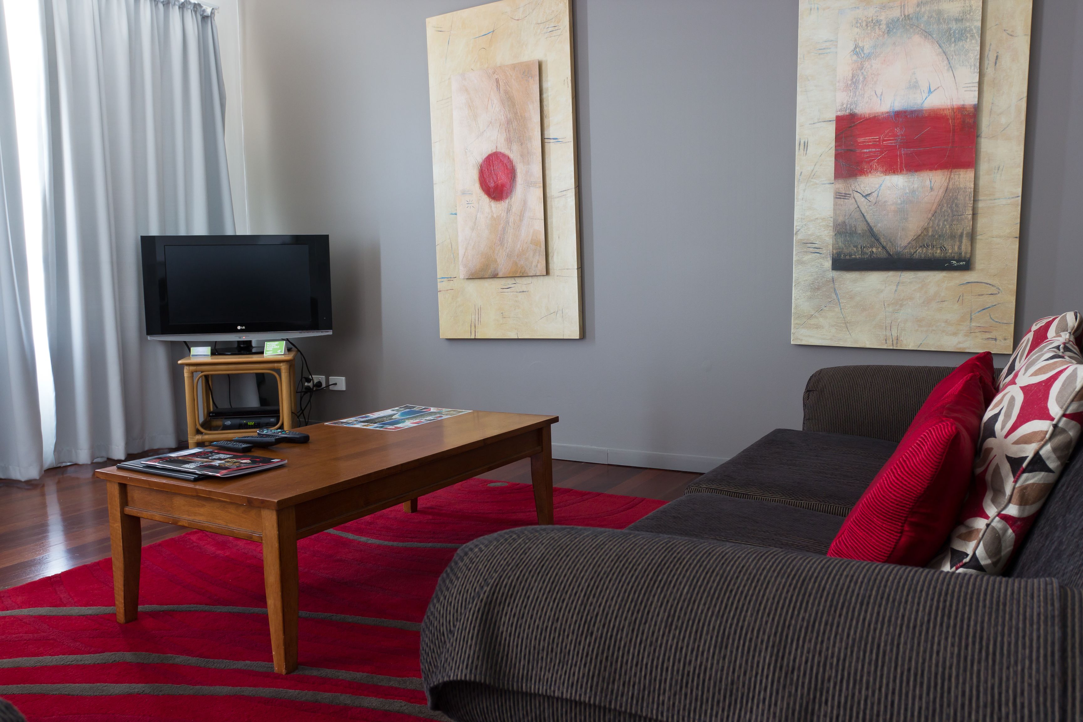 A living room with a flat-screen TV on a wooden stand, a red rug, a grey sofa with red and patterned cushions, and two abstract paintings on the wall.
