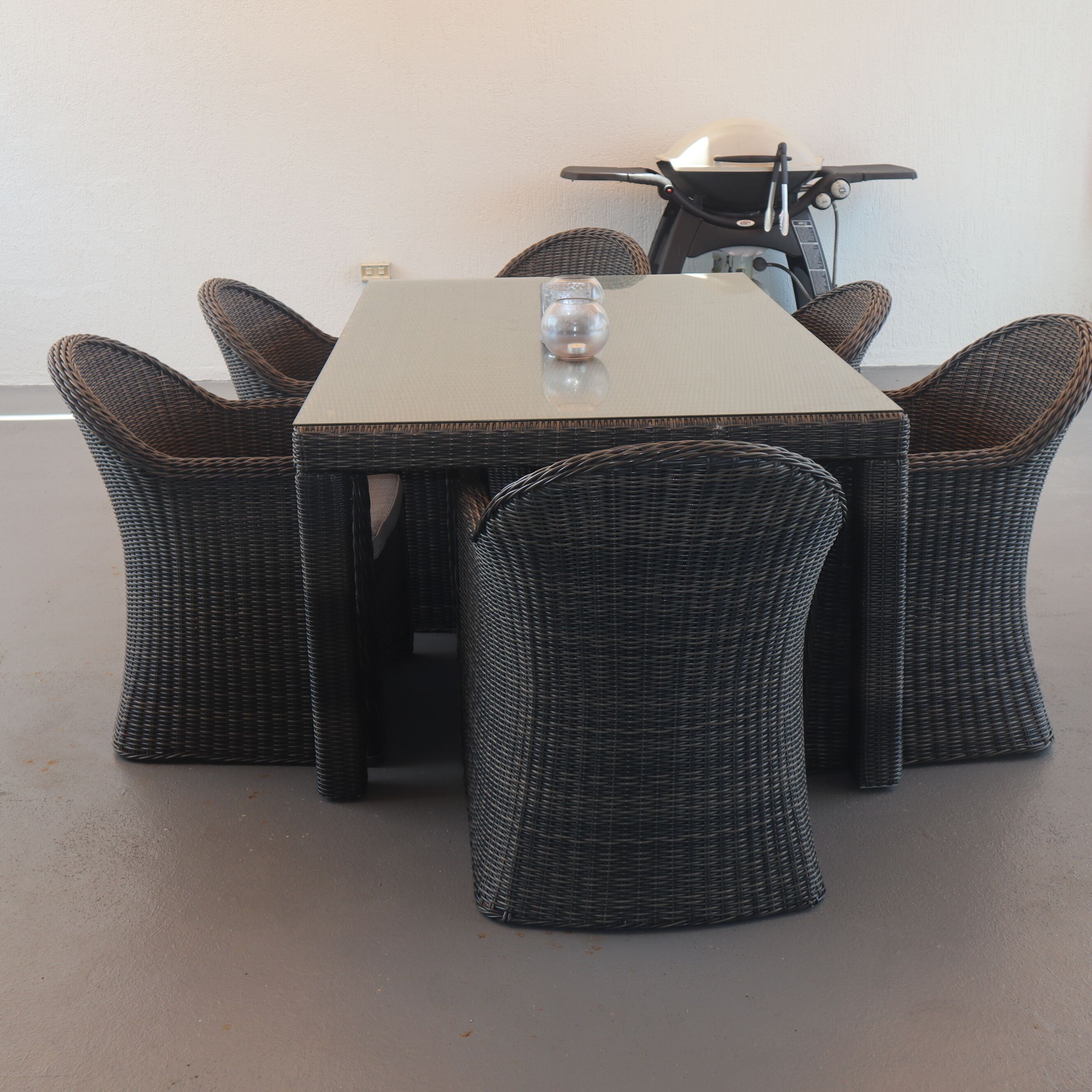 A dining table with six wicker chairs, a glass on the table, and a bicycle in the background.