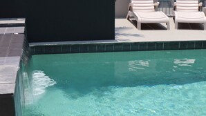 Una piscina cristalina con borde de azulejos y algunas sillas de descanso al fondo.