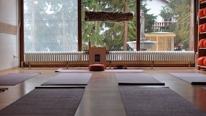 Un estudio de yoga con colchonetas dispuestas en filas, un banco de madera, un radiador y vistas a árboles y una fuente al exterior.