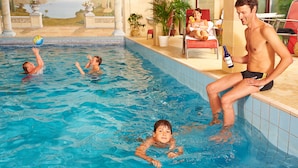 Una piscina con gente en el agua, un hombre sentado en el borde sosteniendo una botella y una mujer en una silla al fondo.