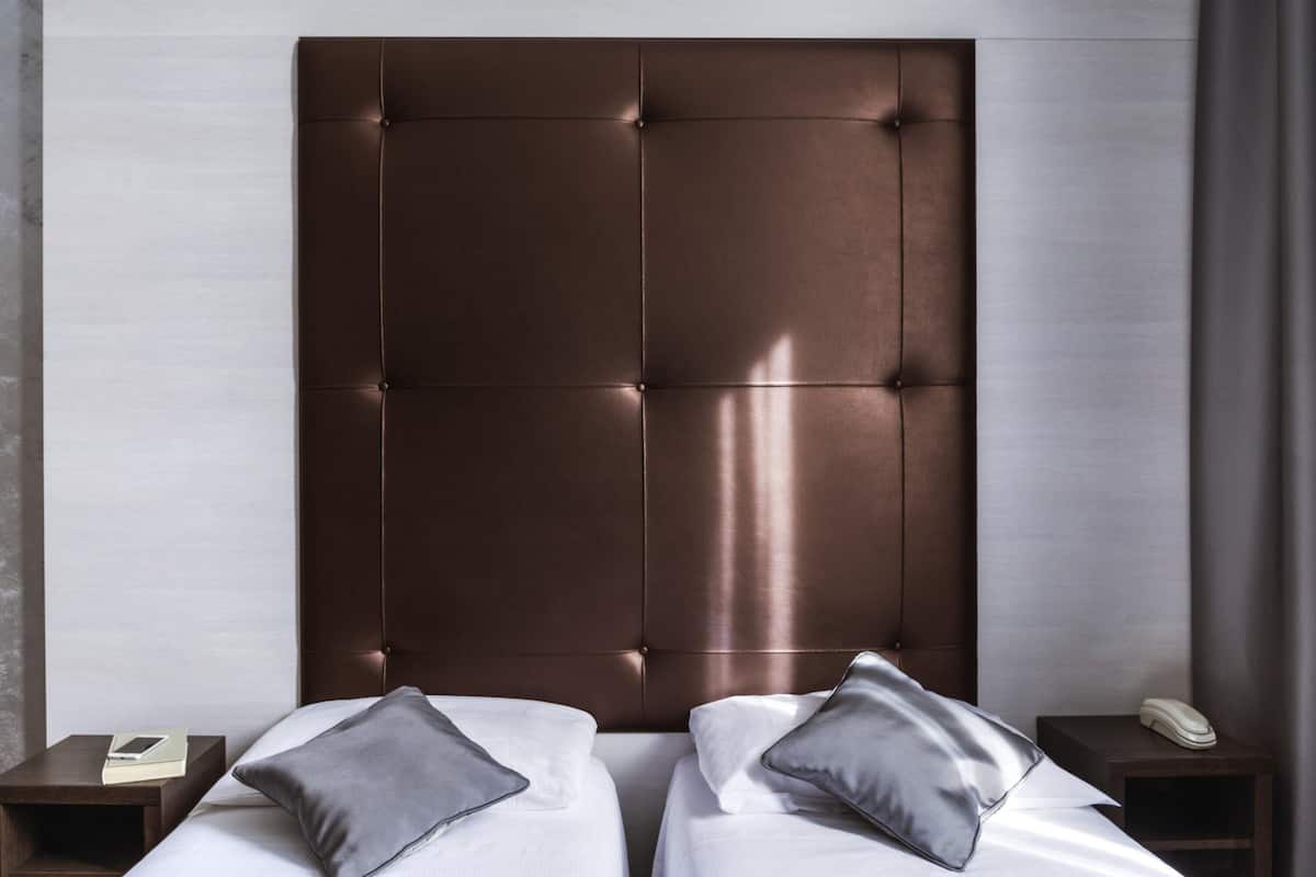 A hotel room with a large, tufted headboard, two beds with white linens, two grey pillows, a bedside table with a phone, and a nightstand with a book.