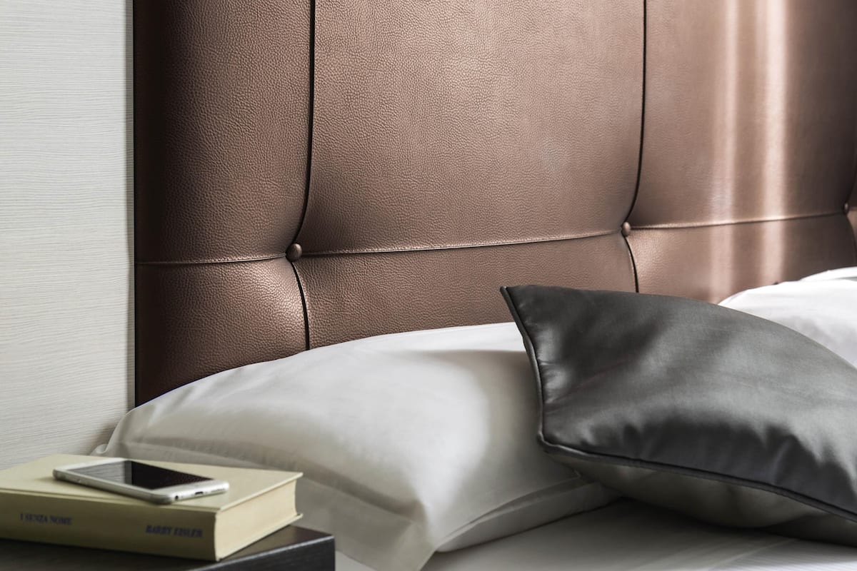Un lit bien fait, avec une tête de lit marron, un oreiller et une table de chevet sur laquelle se trouvent un livre et un téléphone intelligent.